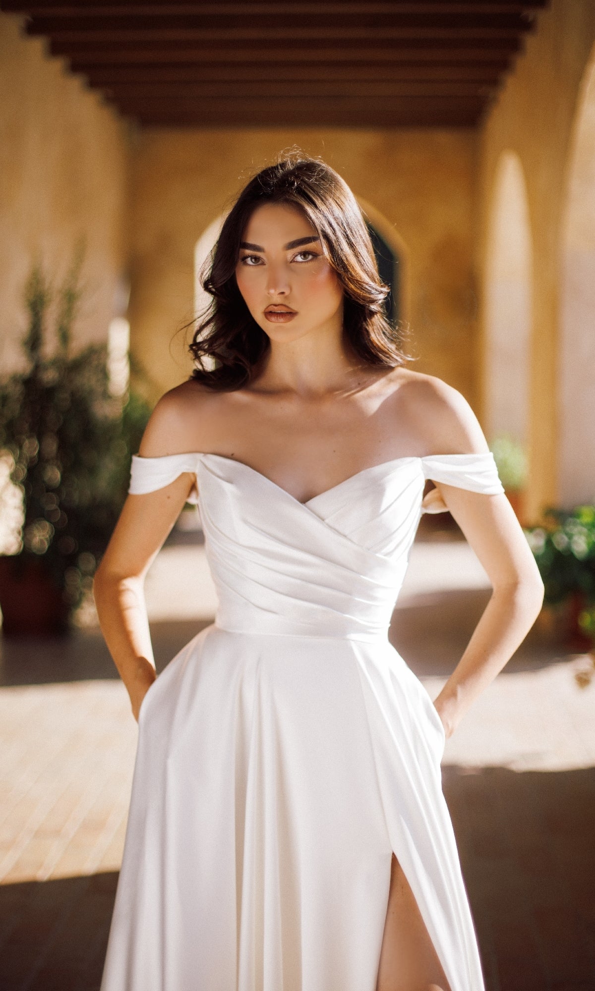 A woman in the Tina Holly TE565 Long Formal Dress stands in a sunlit corridor with her hands in the dress pockets, looking directly at the camera.